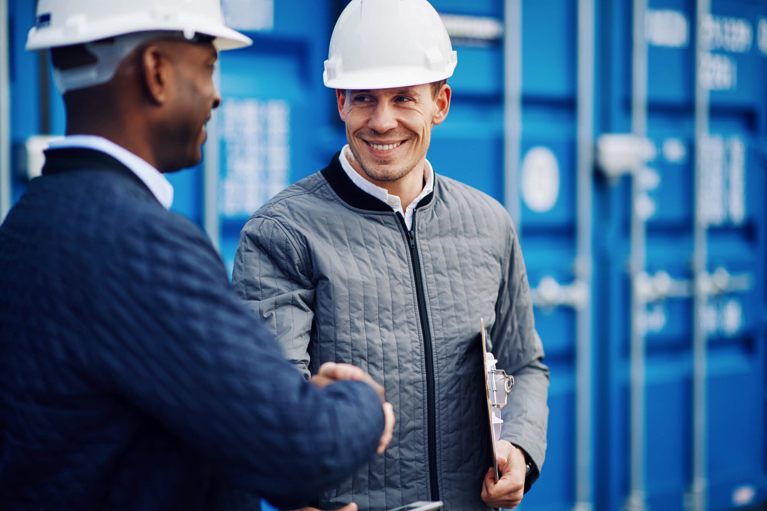 Two smiling engineers wearing hardhats standing by freight containers on a commercial dock shaking hands together